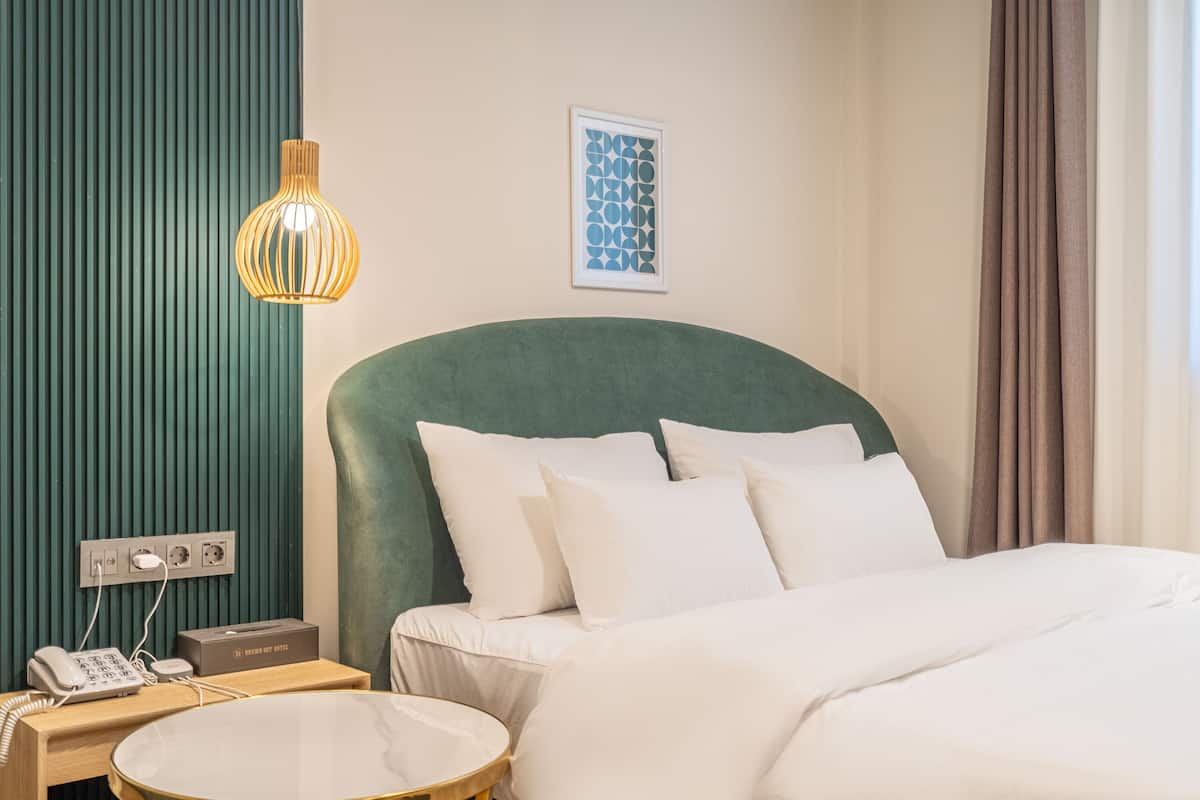 Una habitación de hotel con cabecera verde, ropa de cama blanca, mesita de noche con lámpara, teléfono y un cuadro en la pared.