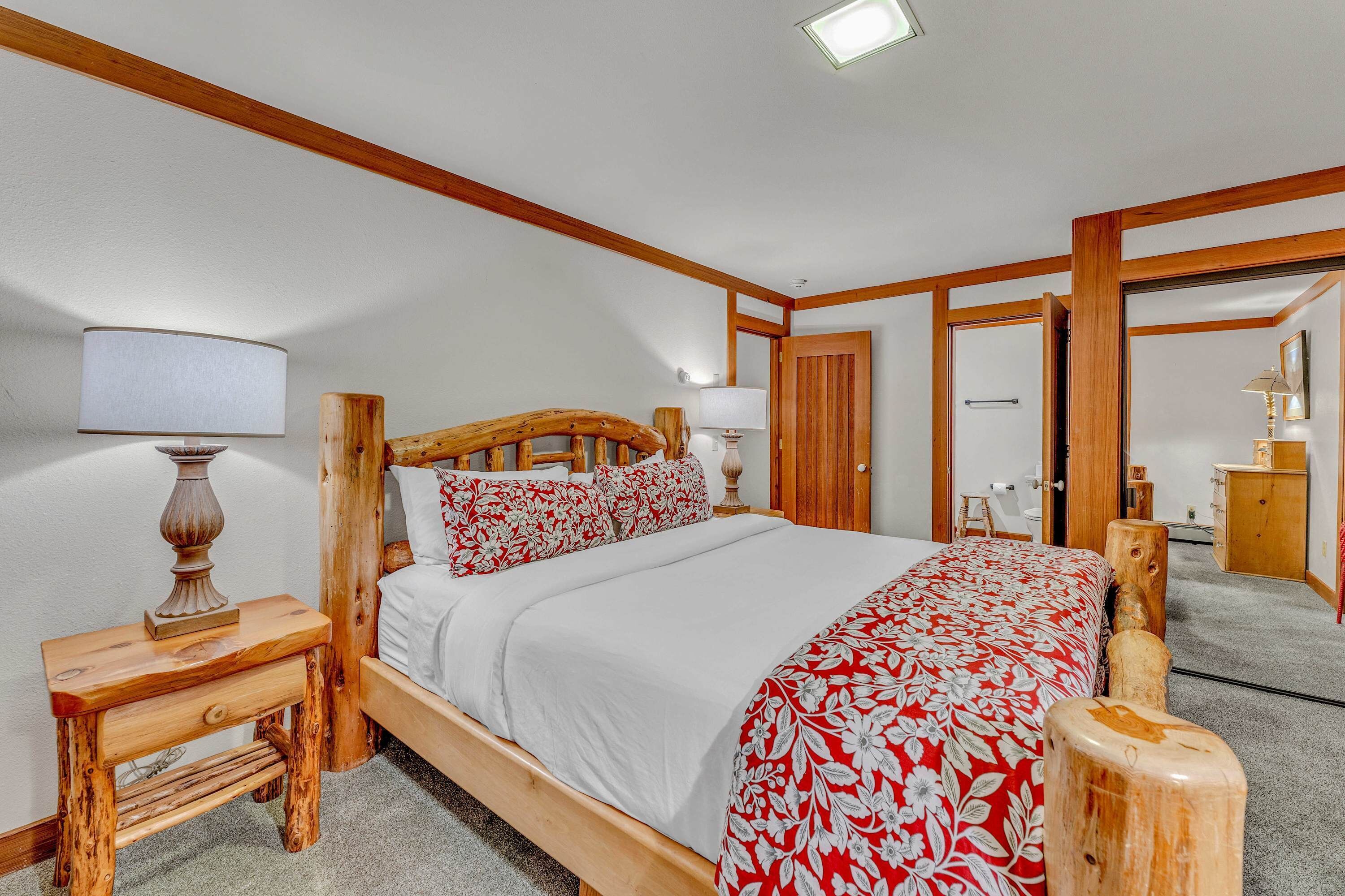 Uma cama com estrutura de madeira, roupa de cama branca e padrões florais vermelhos, um criado-mudo com luminária e cabeceira de madeira.
