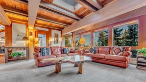 Amplia sala de estar con una gran mesa de centro de madera, sofás de cuero y ventanas amplias que ofrecen vistas a árboles.
