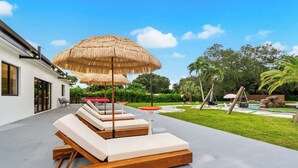 A modern outdoor lounge area with wooden loungers and thatched umbrellas, surrounded by greenery and a swimming pool. - One and Only Lazy River Pool Villa (Miami)