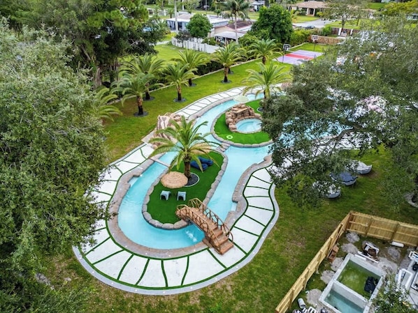 Relax by the winding pool with a central water slide and small waterfall, surrounded by a lush lawn and palm trees.