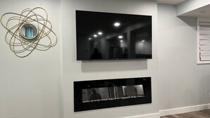 Een moderne woonkamer met een flatscreen televisie aan de muur, een open haard eronder en een decoratief metalen beeld links.
