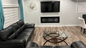 Een moderne woonkamer met een zwarte leren bank, een glazen salontafel, een flatscreen televisie aan de wand en een open haard eronder.