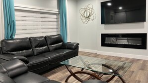 Een moderne woonkamer met een leren zwarte bank, een glazen salontafel, een flatscreen televisie aan de muur en een haard eronder.