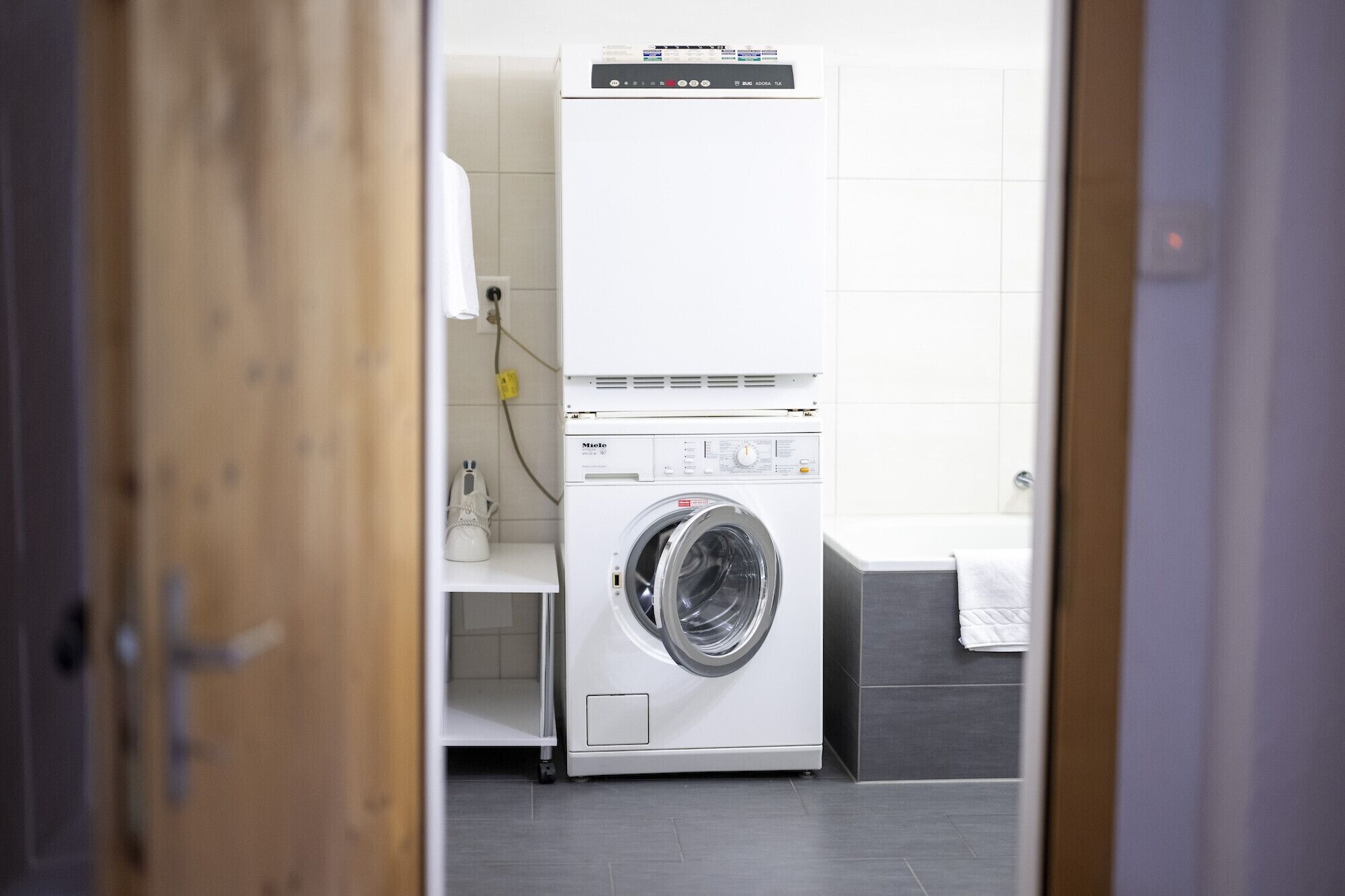 A compact laundry room with a front-loading washing machine and dryer, a small shelf with a white object, and a tiled wall.