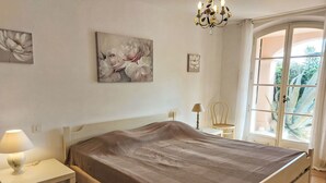Un dormitorio con cama, mesita de noche, lámpara, silla y ventana con vista a follaje.