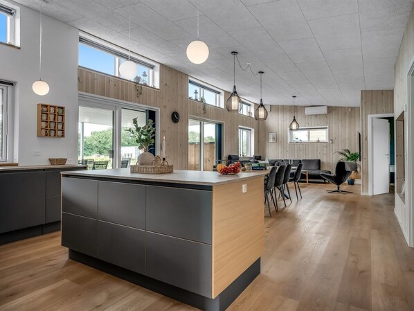 A modern kitchen with a central island, wooden panel walls, large windows, and pendant lighting.