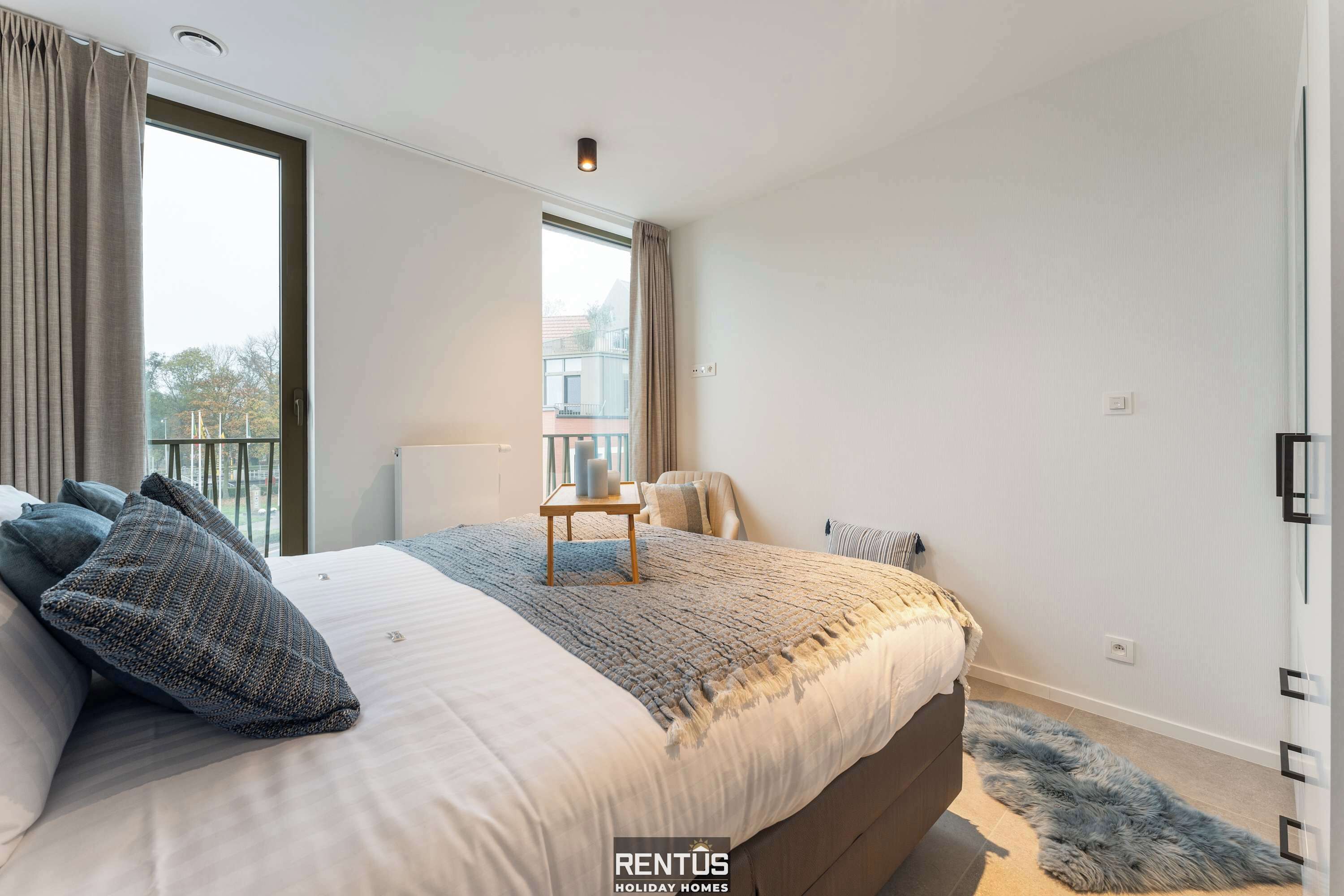 A bedroom with a large bed, a small table, a chair, and a view of the outside.