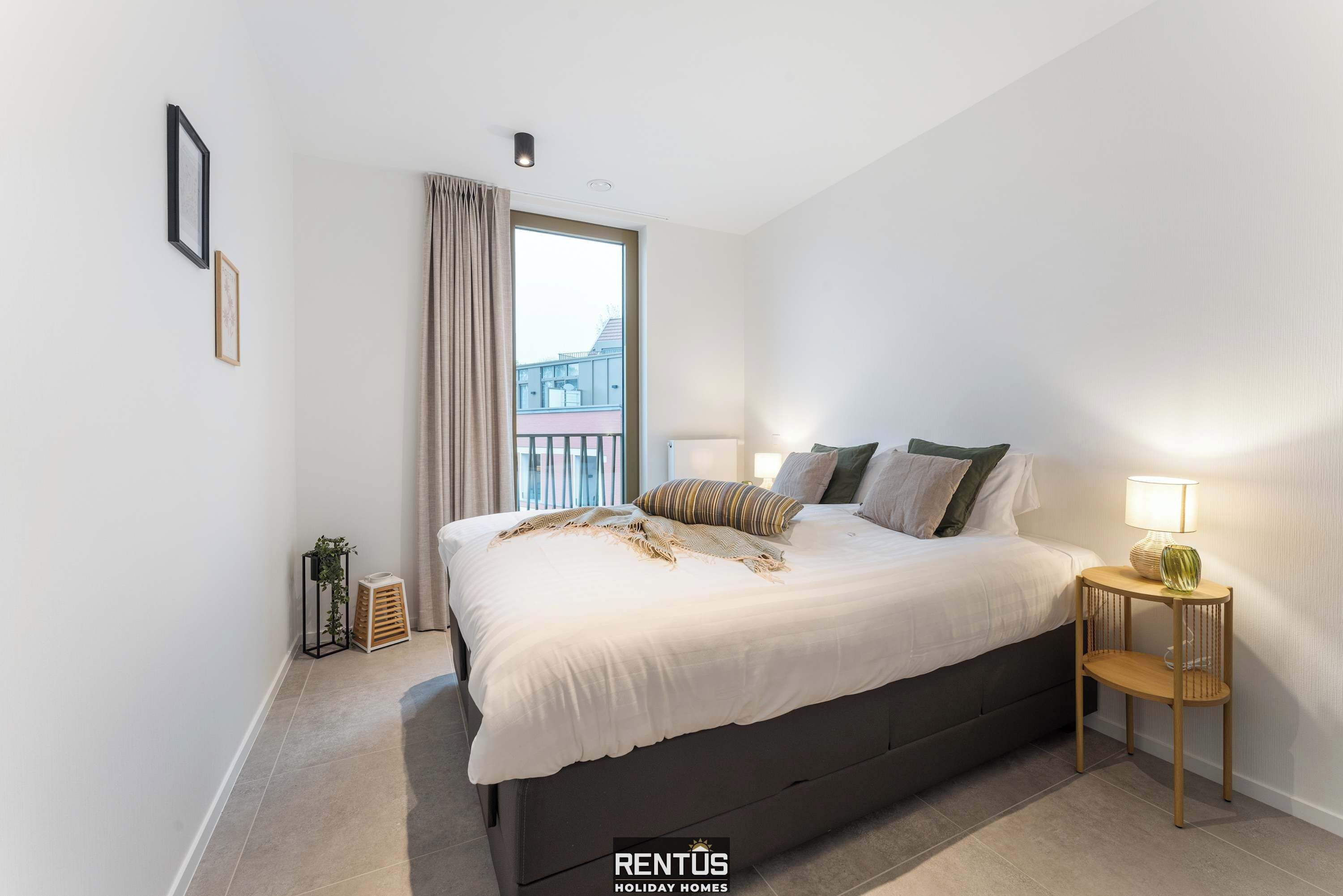 A modern bedroom with a large bed, a bedside table with a lamp, and a view of buildings through the window.
