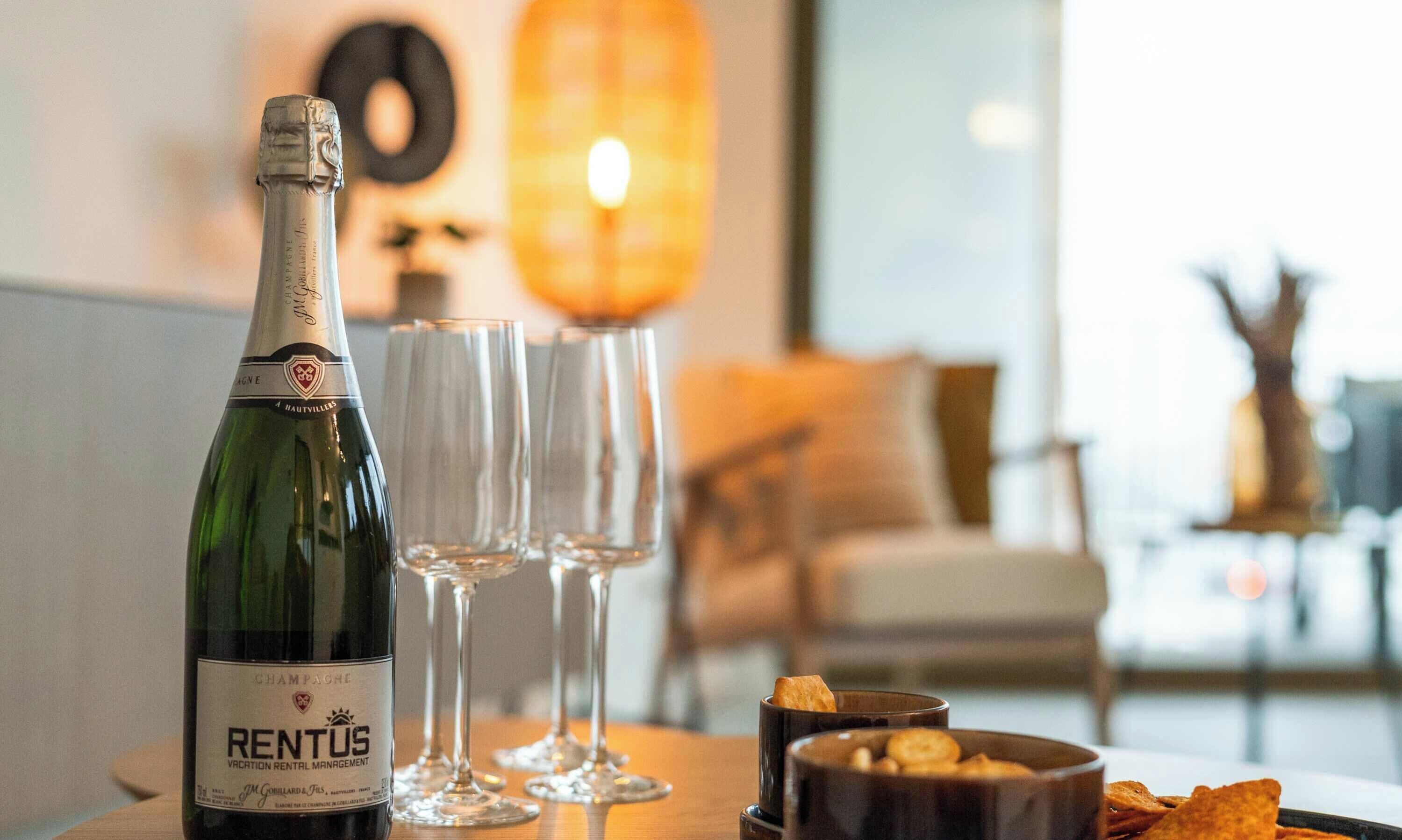 Eine Flasche Rientus Champagner, zwei leere Weingläser und eine Schale mit Snacks auf einem Tisch in einem gut beleuchteten Raum mit modernem Interieur.