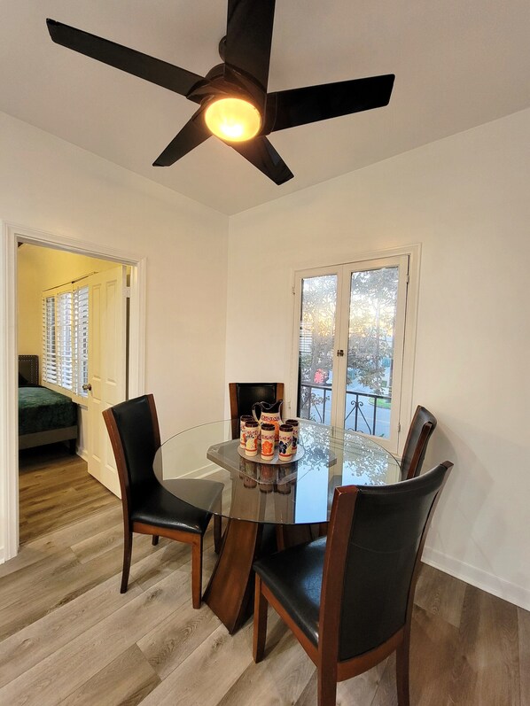 A dining area with a glass table, four chairs, a ceiling fan, and a view of the outdoors through a glass door.