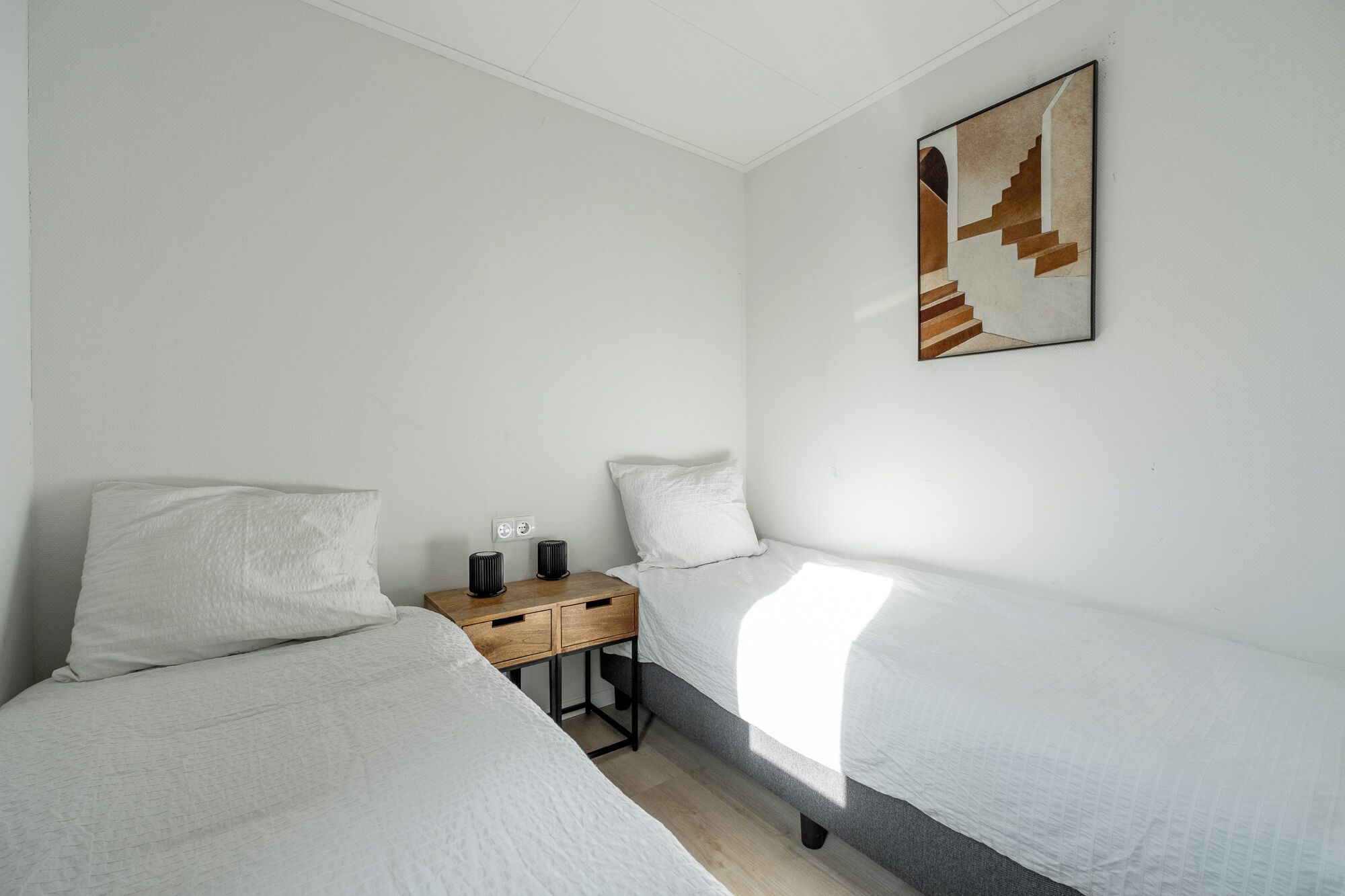 Een slaapkamer met twee enkele bedden, een houten nachtkastje, een kunstwerk aan de muur en een raam dat natuurlijk licht binnenlaat.