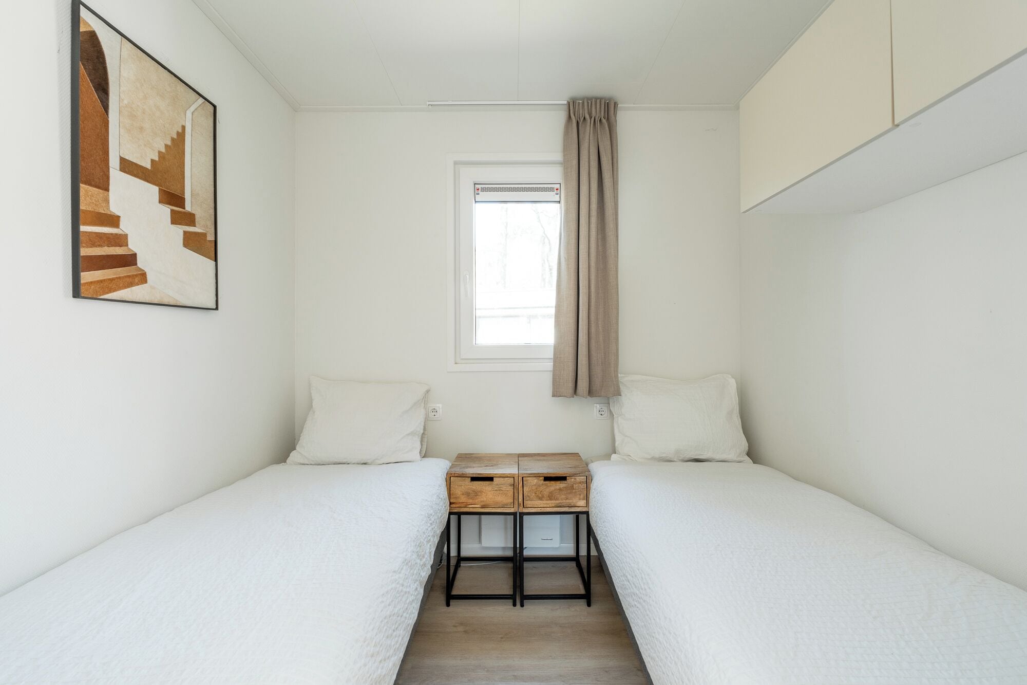 Une petite pièce blanche, composée d’une seule pièce avec deux lits, une fenêtre avec des rideaux, un climatiseur fixé au mur et un tableau encadré.