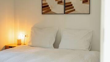Una cama bien tendida con ropa de cama blanca, una lámpara de noche y una obra de arte abstracta colgada en la pared.