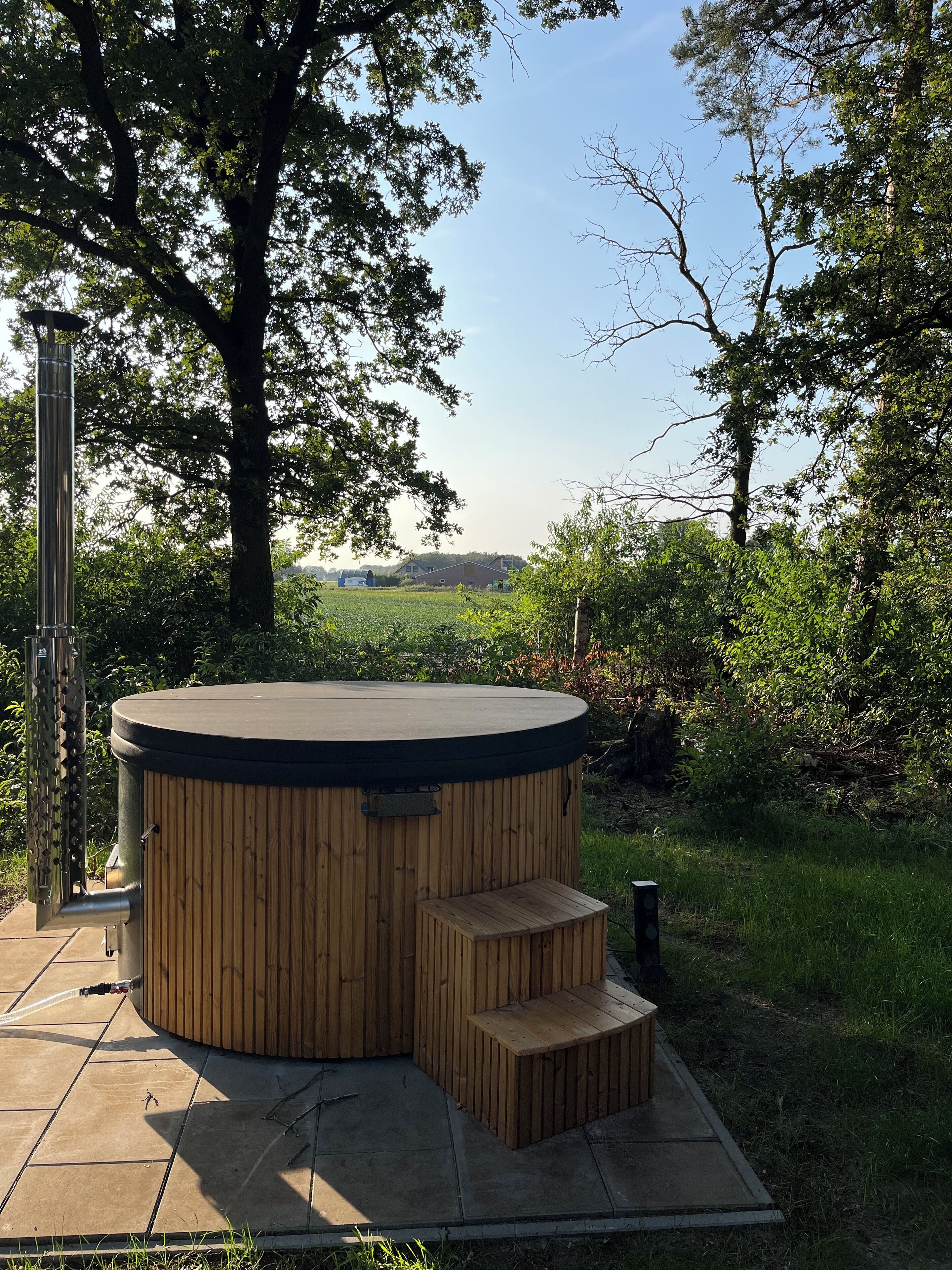 Un jacuzzi con revestimiento de madera y un pequeño banco de madera, rodeado de árboles y un área con césped.