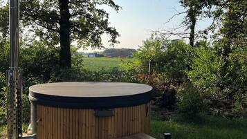 Un jacuzzi con revestimiento de madera y un pequeño banco de madera, rodeado de árboles y un área con césped.