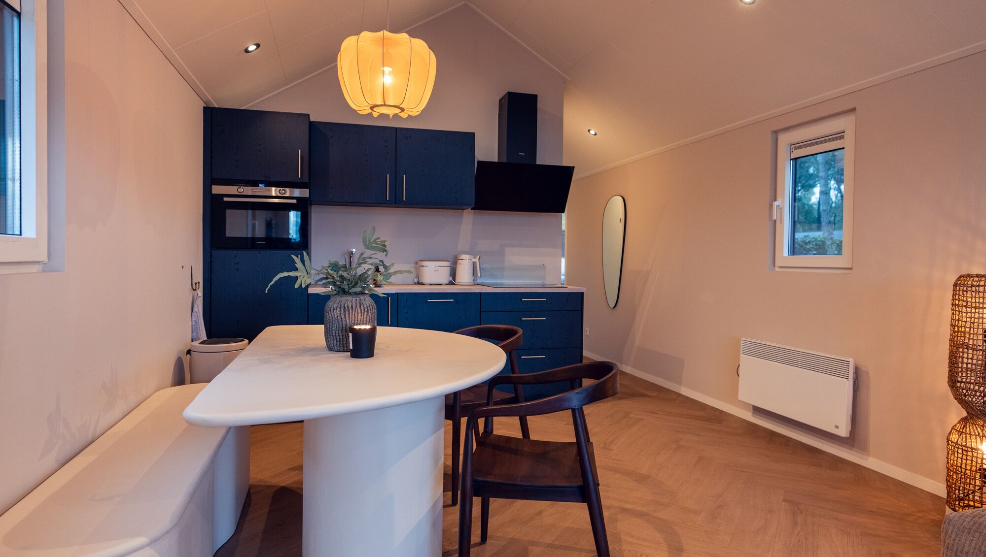 Een moderne keuken met donkerblauwe kastjes, een wit aanrechtblad en een centraal eiland. Er is een inbouwoven en magnetron, een hanglamp en een raam met uitzicht op bomen.