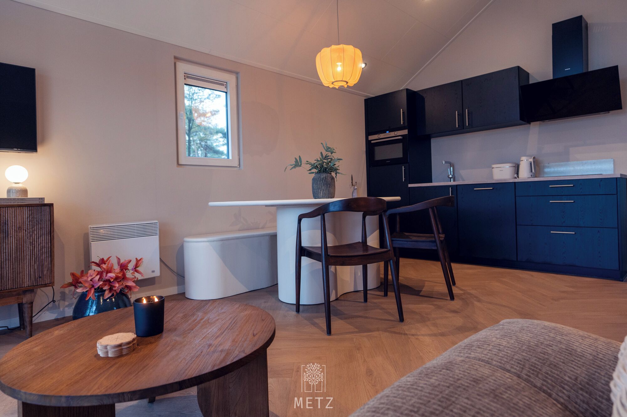 Een moderne keuken met donkere kastjes, een wit aanrechtblad en een inbouwoven. Er is een eethoek met een houten tafel en twee stoelen. Op de tafel staan een potplant en een kaars.