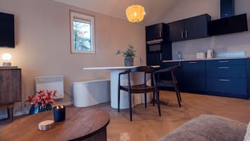 Een moderne keuken met donkere kastjes, een wit aanrechtblad en een inbouwoven. Er is een eethoek met een houten tafel en twee stoelen. Op de tafel staan een potplant en een kaars.
