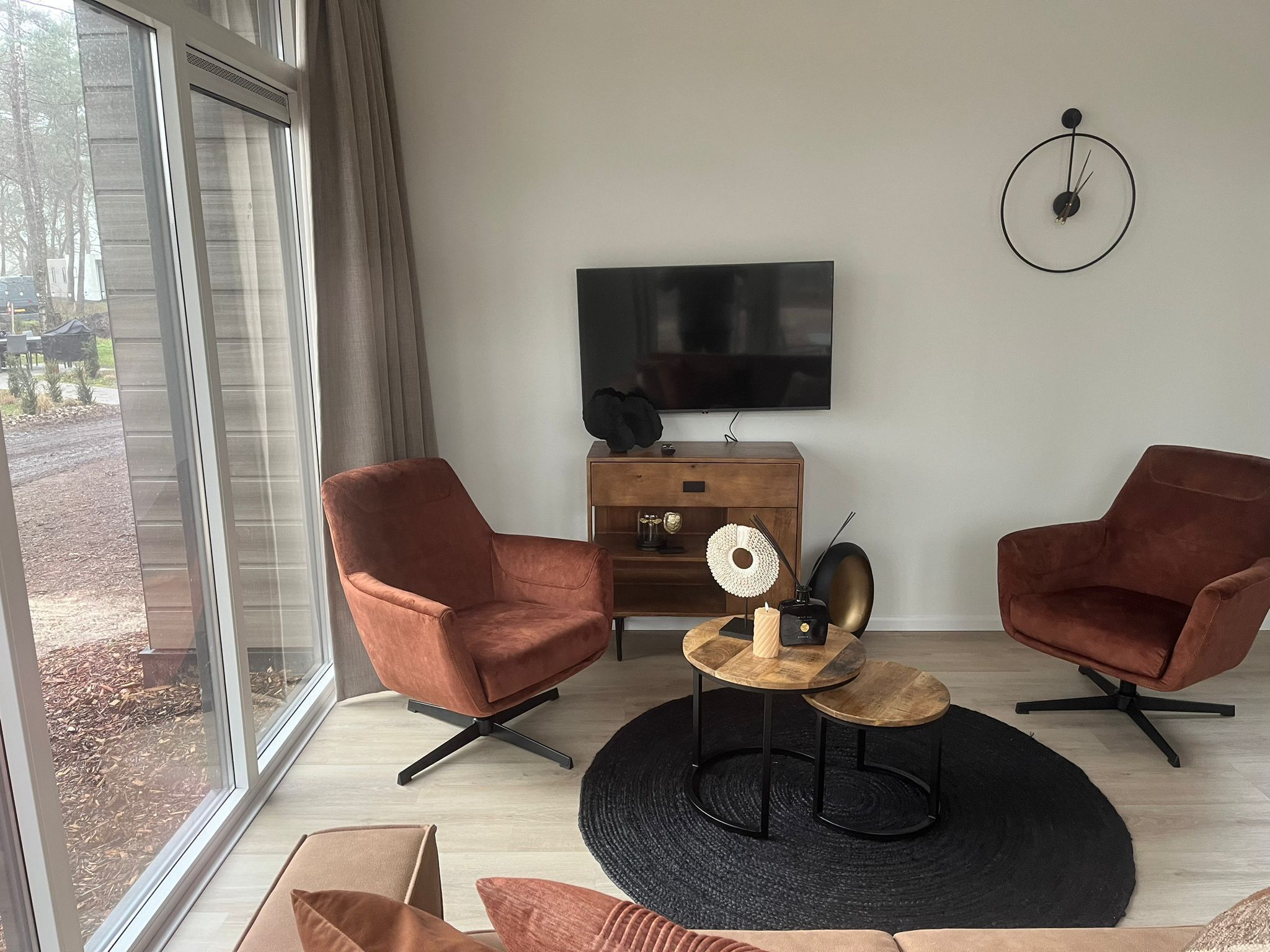 A modern living room with two brown armchairs, a wooden coffee table, a black rug, and a TV mounted on the wall.