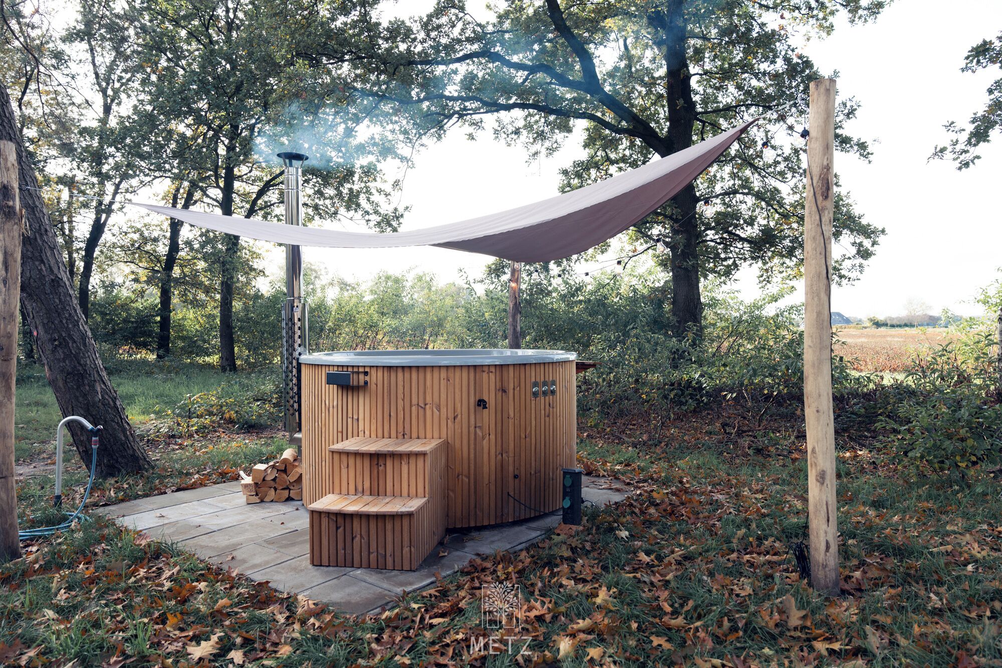 A wooden hot tub with a metal frame, surrounded by trees and a hammock.