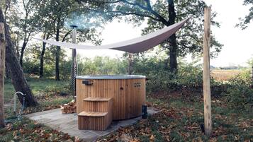 A wooden hot tub with a metal frame, surrounded by trees and a hammock.