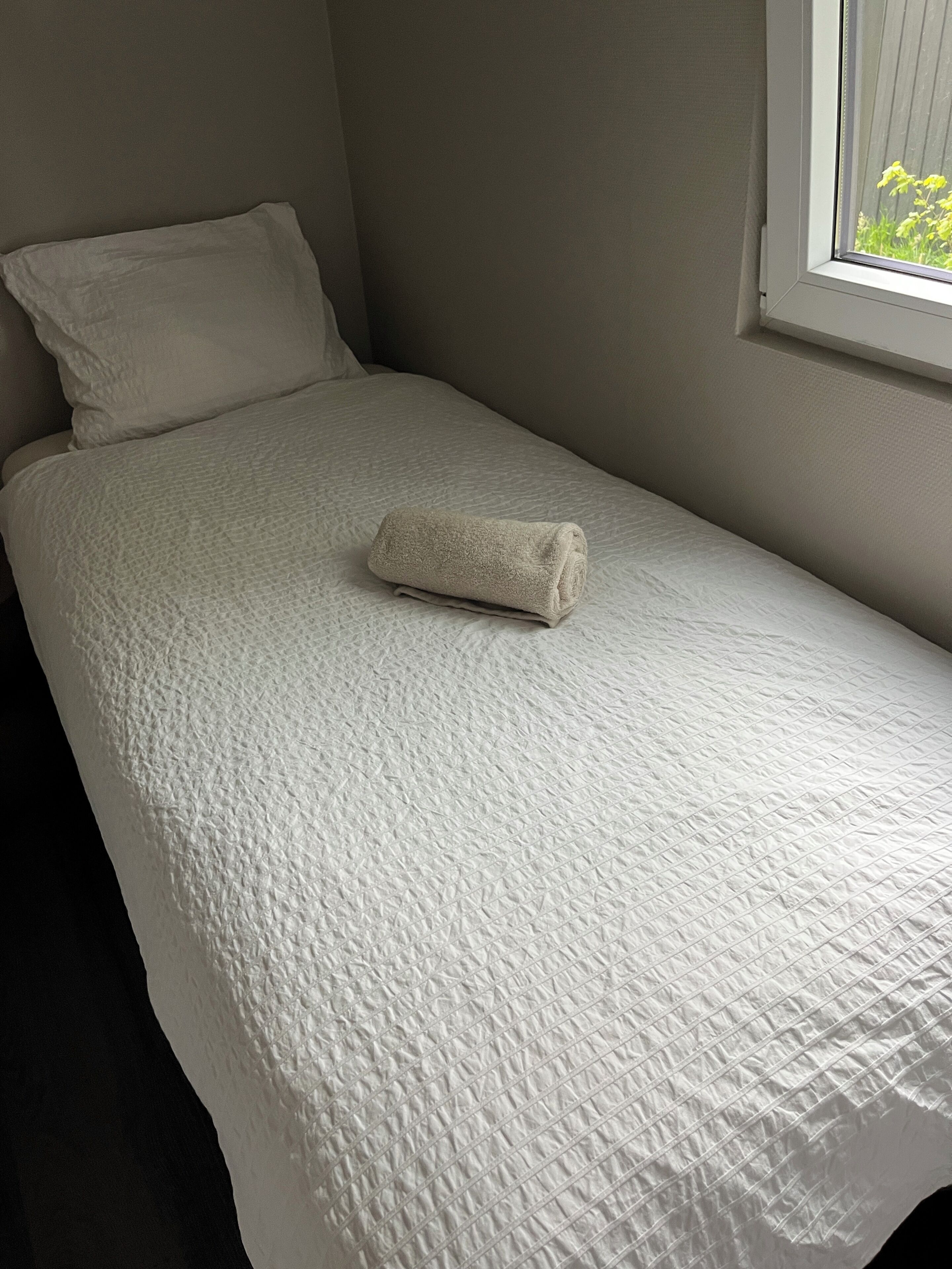 A single bed with a rolled towel, plain walls, and a window with a view of greenery.