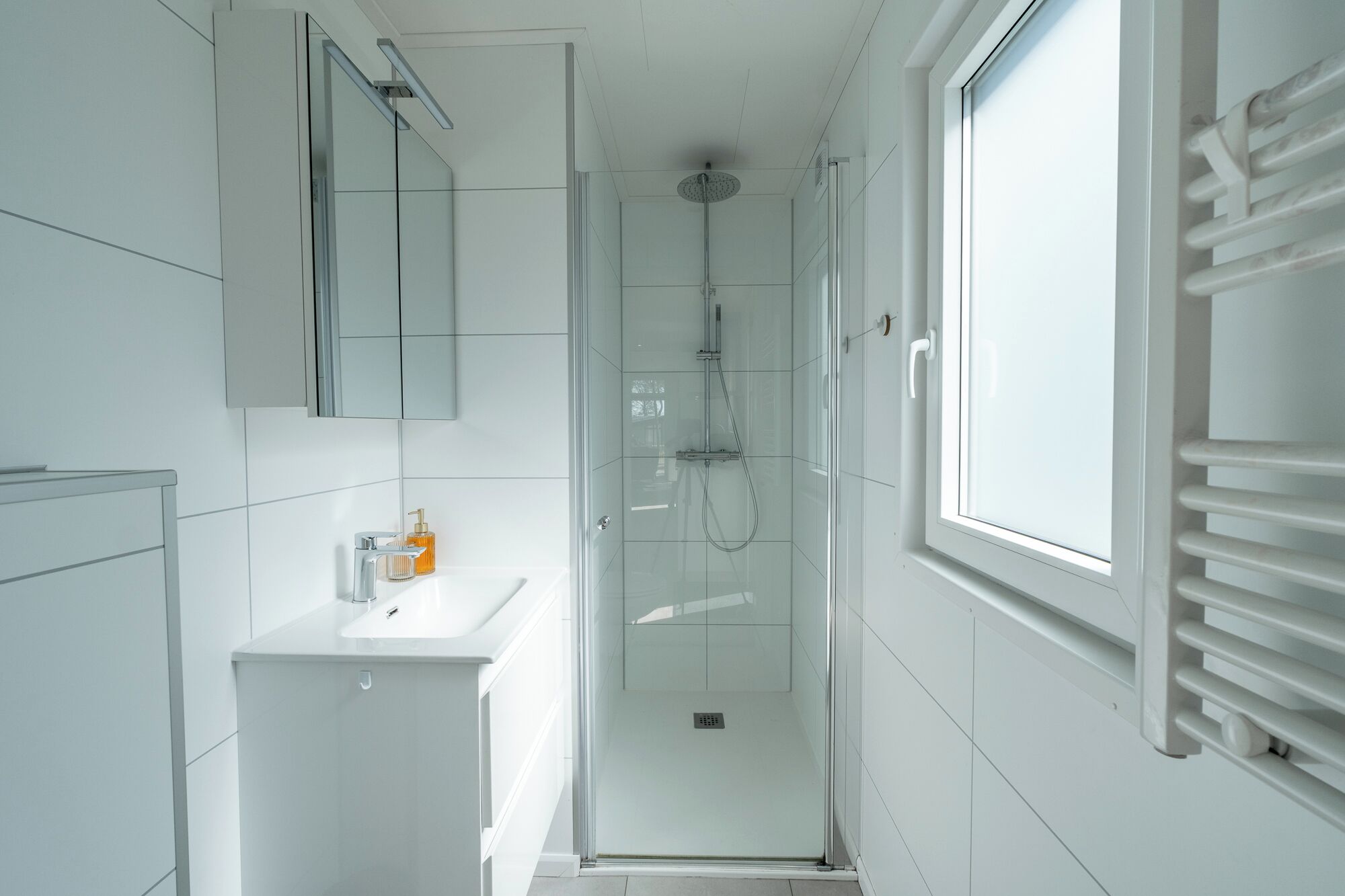 Un baño moderno con ducha de vidrio, un lavamanos blanco y un ventanal amplio.