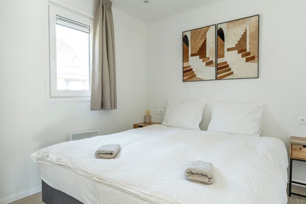 A bedroom with a bed, two pillows, a nightstand, a lamp, a window with curtains, and two wall-mounted paintings. - Wellness Lodge Hottub (Haarle)