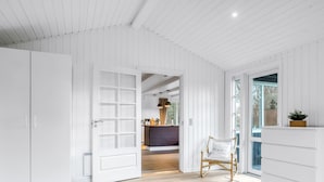 Een moderne slaapkamer met een groot bed, een houten vloer, witte wanden en uitzicht door een geopende deur naar een eetgedeelte.