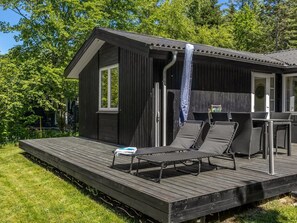 Eine Holzterrasse mit Liegestühlen und einem kleinen Tisch, ein überdachter Außenbereich mit Wasserspiel und ein dunkles Holzgebäude mit Fenster und Tür.