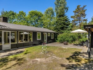 A small building with large windows, a patio area with outdoor furniture, and a garden with trees.