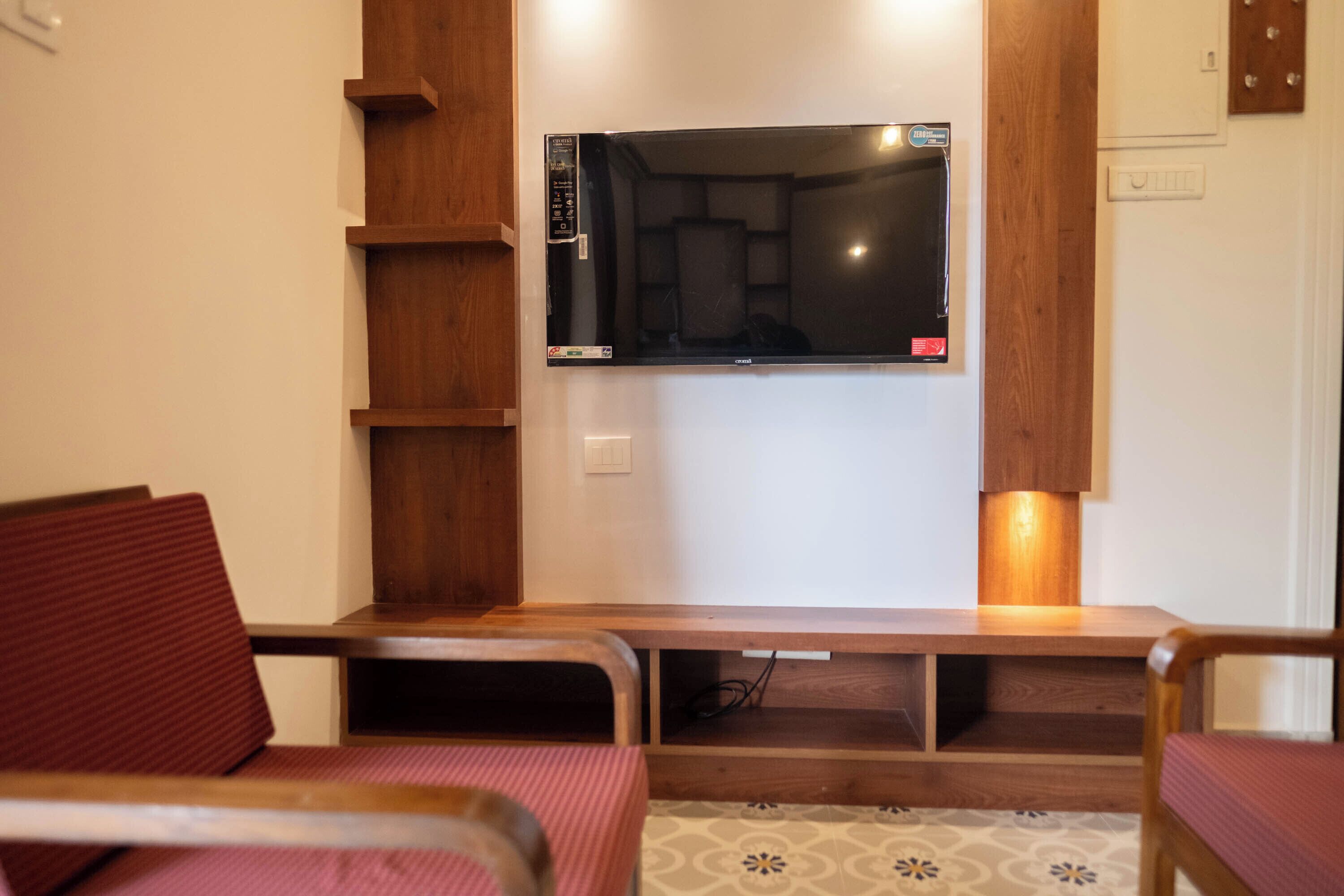 A living room with a flat-screen TV mounted on the wall, wooden shelving, and a red cushioned chair.