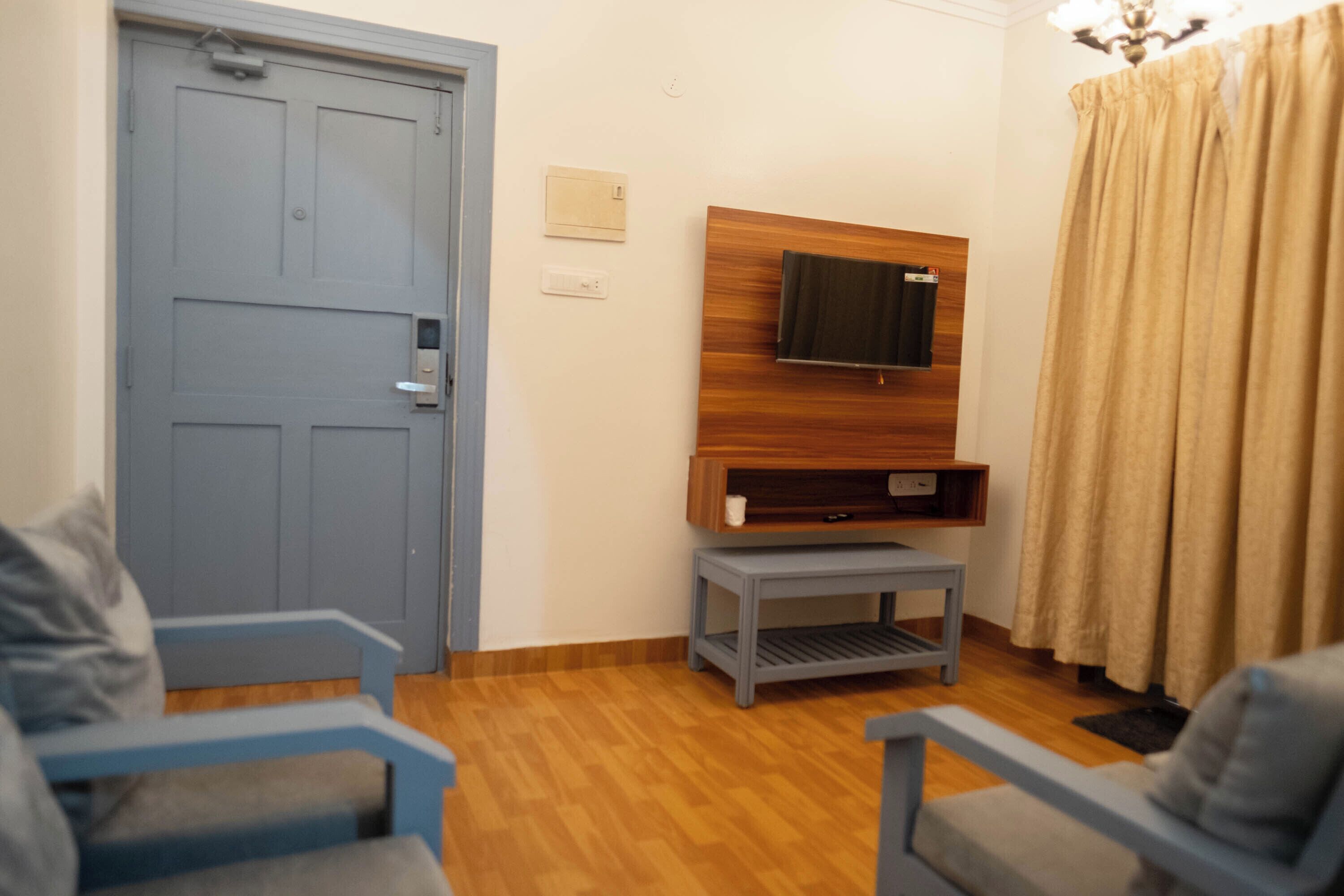 Ein Zimmer mit einer blauen Tür, einem an einer Holzplatte montierten Fernseher, einem kleinen grauen Tisch und einem grauen Sofa.