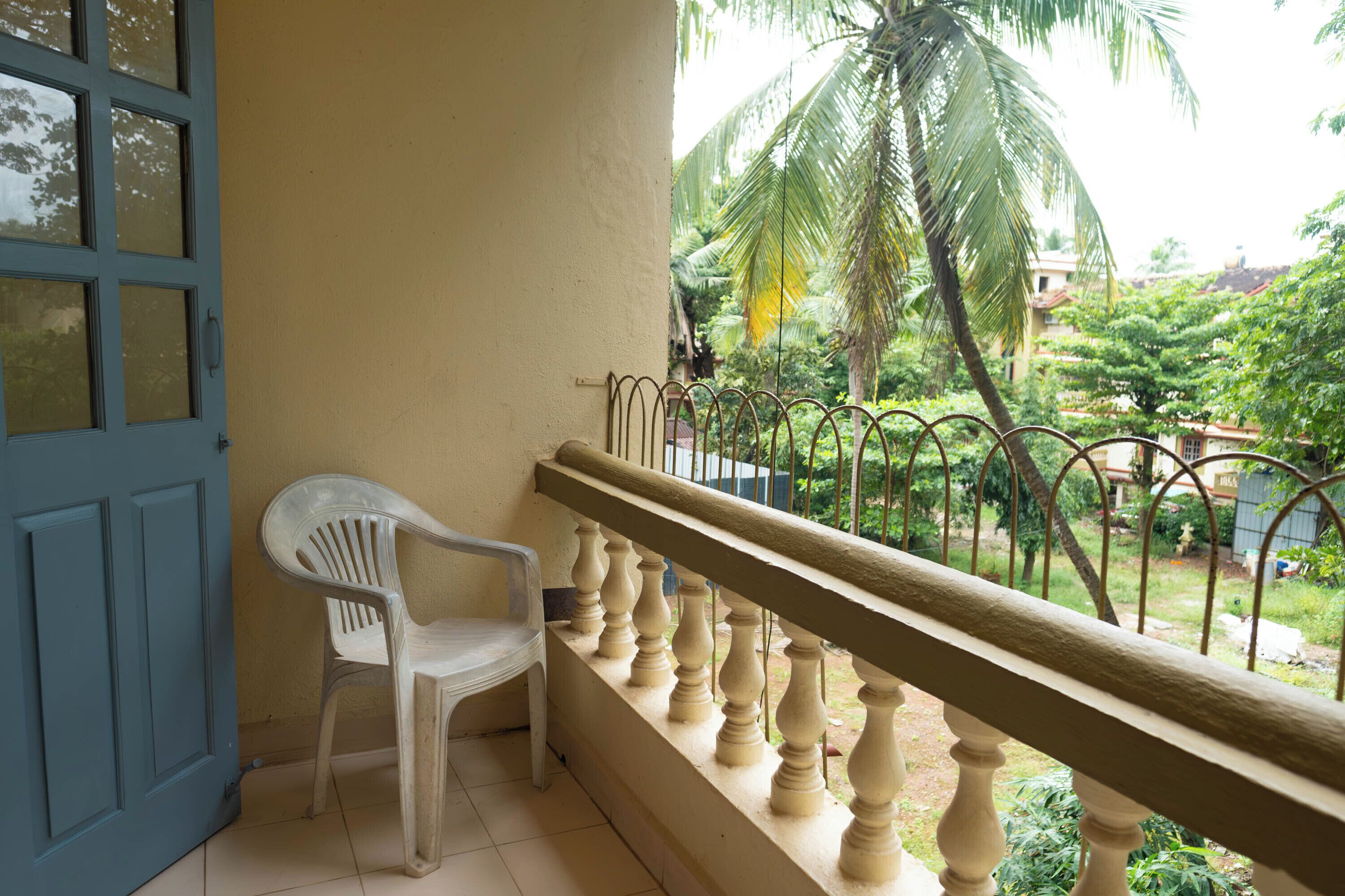 A balcony with a white plastic chair, a blue door, and a view of a lush garden with palm trees.