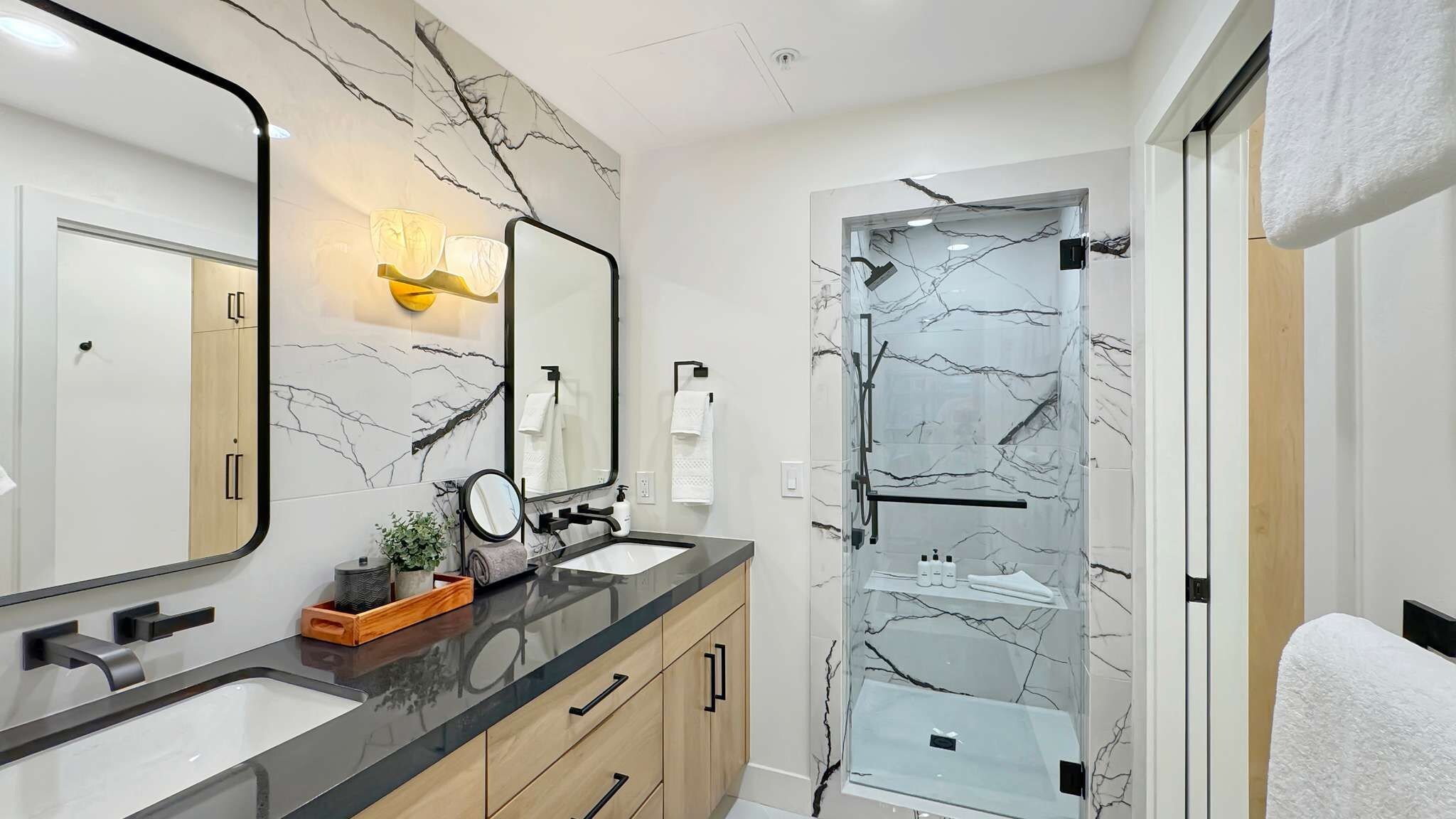 A modern bathroom with marble walls, a dual sink vanity, and a glass shower enclosure.