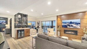 A modern living room with a fireplace, a kitchen with a glass backsplash, and a large window offering a view of the mountains.
