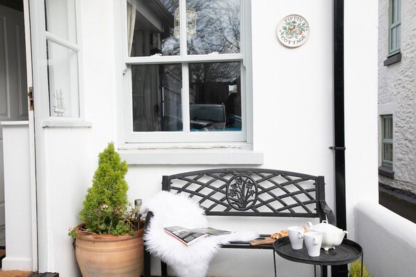 A white cottage exterior with a black metal bench, a small table with cups and a teapot, and a potted plant.