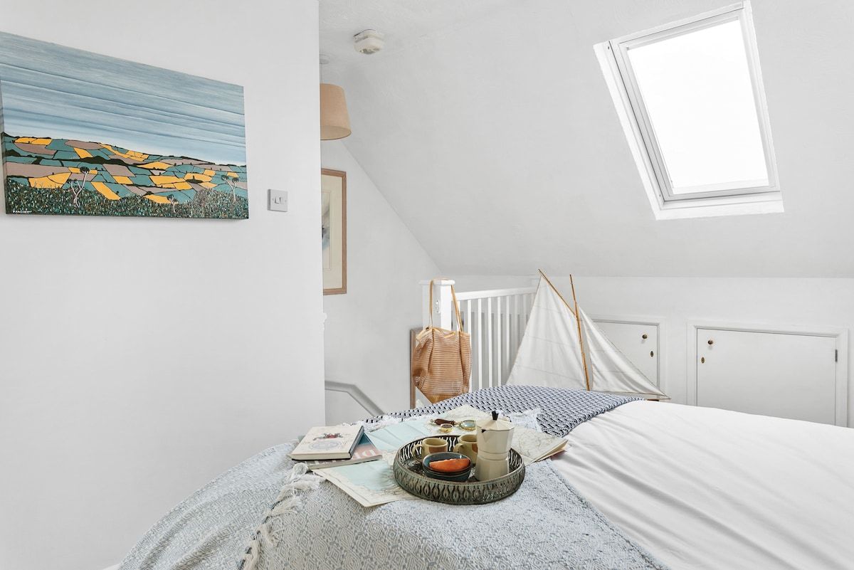 A bedroom with a bed, a window, a painting, and a bedside table with items.