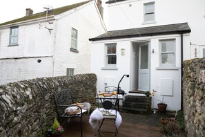 Una casa blanca con un muro de piedra, un pequeño espacio al aire libre con sillas y mesa de metal, y una puerta que da a la entrada.