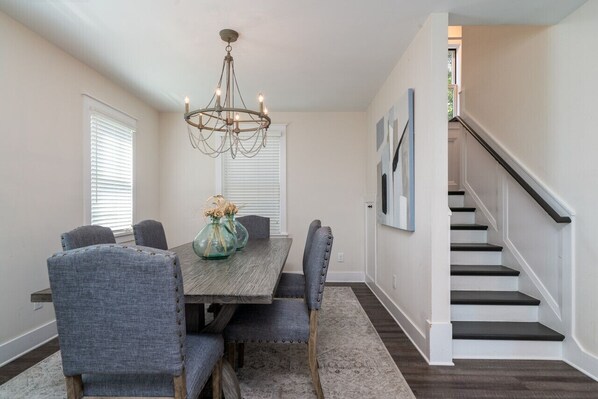 A dining area with a wooden table and six upholstered chairs, a chandelier, and a staircase leading to another level.