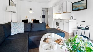 Una sala de estar moderna con un sofá oscuro, un comedor con una mesa preparada para dos personas, y una cocina con armarios blancos y un televisor de pantalla plana.