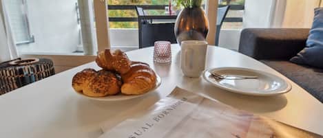 A table with croissants, a vase with flowers, a cup, and a plate with a spoon. A menu titled "VISUAL BOOKHOLM" is on the table.