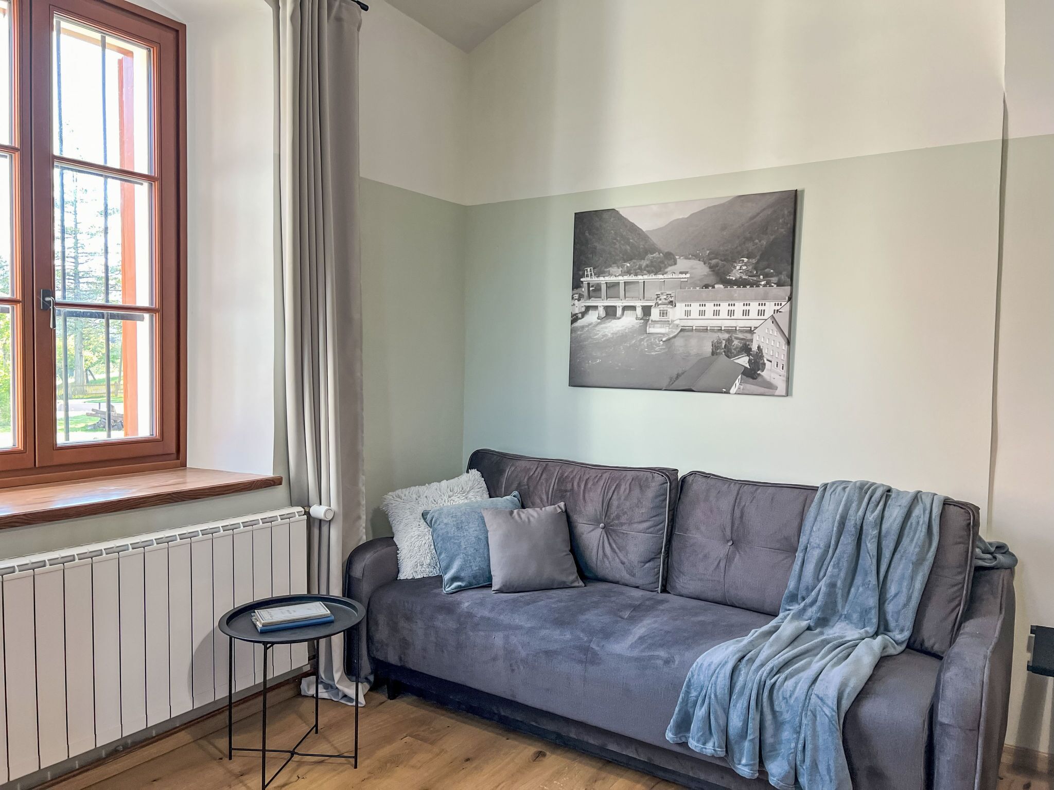 A cozy room with a grey sofa, a small side table, a window with curtains, and a black and white photograph on the wall.