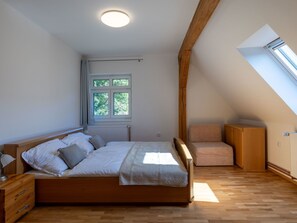 A bedroom with a wooden bed, a window with curtains, a radiator, a wooden chair, and a sloped ceiling with a skylight.