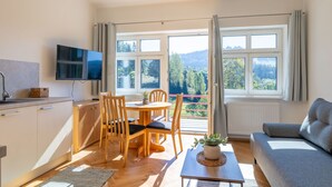 Una sala de estar moderna con un sofá, una mesa de comedor con sillas, un televisor montado en la pared y ventanas amplias con vistas al exterior.