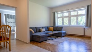 Una sala moderna con un sofá seccional gris, una silla de madera, una mesita y un ventanal grande con cortinas.
