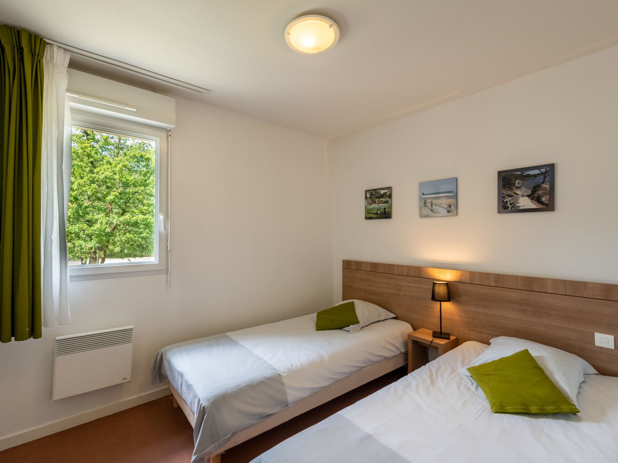 A hotel room with two single beds, a window with curtains, a wall-mounted heater, and three framed pictures.