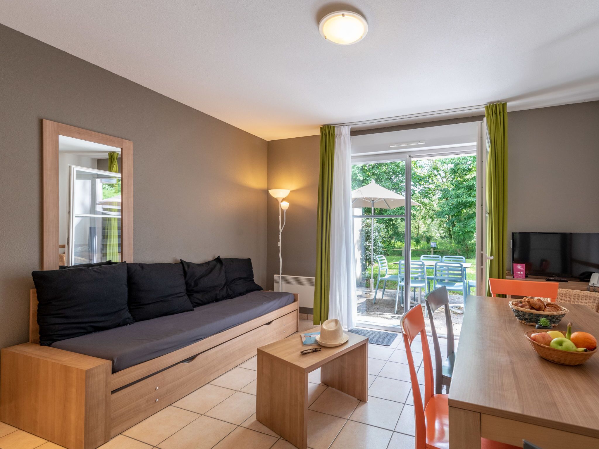 Una sala de estar moderna con sofá, una mesa de comedor y televisor. Hay un ventanal corredero que da a un espacio exterior con sillas y una mesa.
