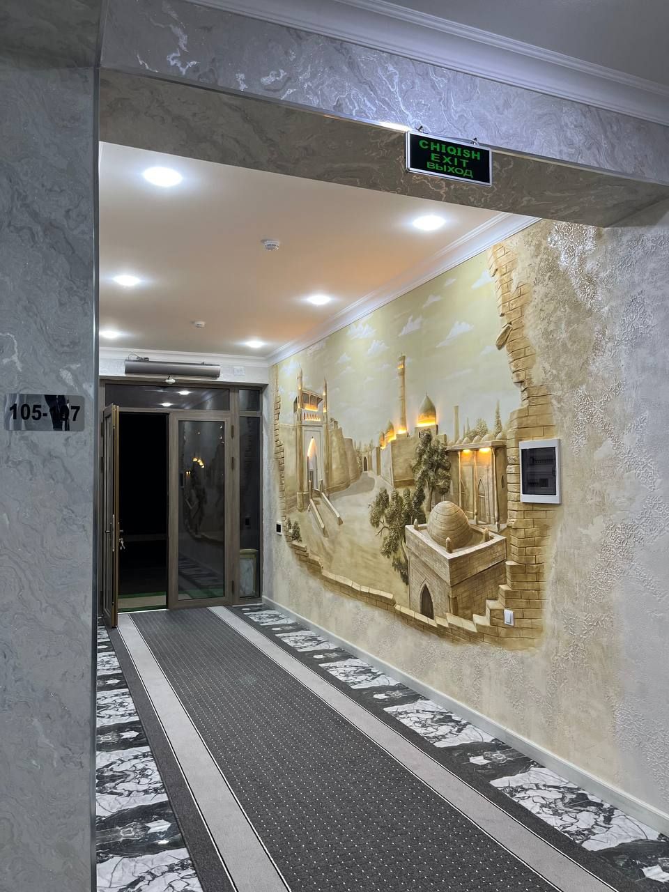 Een hotel lobby met een marmeren vloer, een muurschildering van een historisch tafereel en een bord dat de uitgang aanwijst.